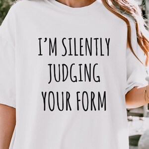 May include: A white short-sleeved t-shirt featuring the black text "I'M SILENTLY JUDGING YOUR FORM" printed in a simple sans-serif font on the chest. The garment has a crew neckline and a loose, relaxed fit, paired with light-colored shorts.