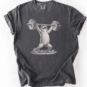May include: Dark gray t-shirt featuring a whimsical illustration of a frog lifting a barbell overhead. The frog is depicted in a vintage, etched style, with the barbell and frog in white against the dark gray fabric. The shirt has a classic crew neck and short sleeves.