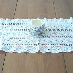 May include: A white lace table runner with a scalloped edge, featuring a repeating geometric pattern. A floral teacup and saucer sit atop the runner. The teacup has a white interior and a handle. The background is a wooden surface.