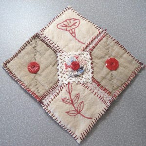 May include: A square fabric coaster featuring a patchwork design with cream and tan panels. It includes red embroidered floral motifs, two red buttons, and a central white lace square topped with a small patterned fabric circle and red button.