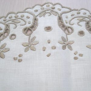 K&ouml;nnte beinhalten: Nahaufnahme eines wei&szlig;en Leinentextils mit aufwendiger Stickerei. Das Design zeigt einen gewellten Rand mit floralen und Rankenmustern in Hellbraun. Kleine, verstreute Stickdetails sorgen f&uuml;r Textur.