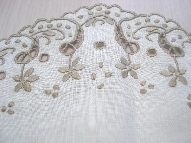 K&ouml;nnte beinhalten: Nahaufnahme eines wei&szlig;en Leinentextils mit aufwendiger Stickerei. Das Design zeigt einen gewellten Rand mit floralen und Rankenmustern in Hellbraun. Kleine, verstreute Stickdetails sorgen f&uuml;r Textur.