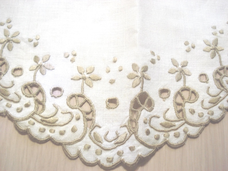 K&ouml;nnte beinhalten: Cremefarbener Stoff mit gesticktem Blumen- und Rankenbord&uuml;ren. Das Design zeigt stilisierte Blumen und dekorative Ausschnitte mit einem gewellten Rand. Die Stickerei ist in einem ged&auml;mpften Goldton gehalten und erzeugt eine zarte und Vintage-&Auml;sthetik.