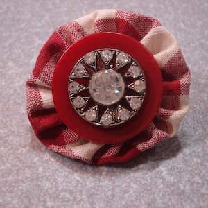 May include: A fabric flower pin with a red and white gingham pattern. The center features a red button with a silver-toned starburst design and a large, clear rhinestone. A decorative accessory.