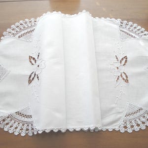 May include: White oval-shaped table runner with a delicate floral cut-out design. The edges are trimmed with a detailed, scalloped lace. The runner is folded, showcasing the intricate craftsmanship and texture of the fabric.