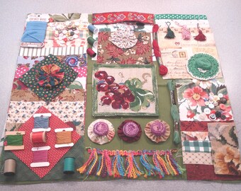 May include: A patchwork of fabric squares in various floral and patterned designs, with spools of thread, buttons, and decorative elements. Includes a framed grape illustration, tassels, and fabric flowers. The color palette is dominated by greens, reds, and blues.