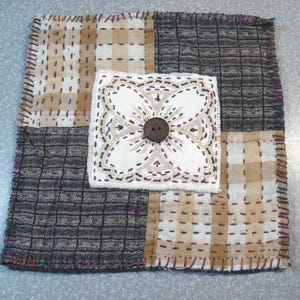 May include: A square textile patch featuring a patchwork design of beige and white checkered fabric alternating with dark gray textured fabric. A central white square panel with intricate floral embroidery and a single dark brown button.