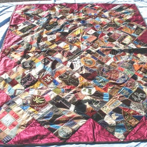 May include: A handmade quilt with a patchwork design. The quilt features a variety of fabric pieces in various colors, including red, brown, blue, and green. The quilt has a burgundy border and is displayed on a light blue surface.