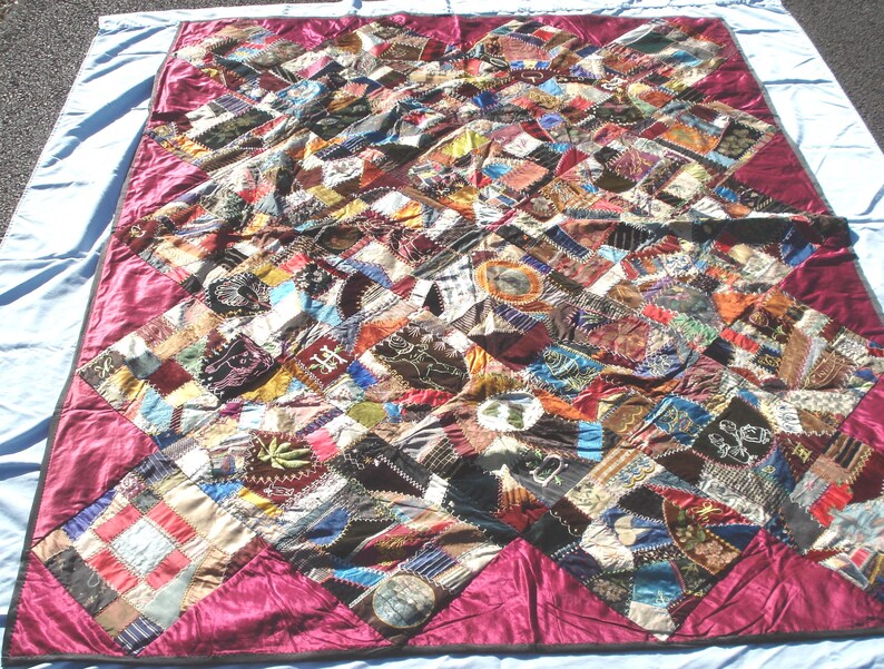 May include: A handmade quilt with a patchwork design. The quilt features a variety of fabric pieces in various colors, including red, brown, blue, and green. The quilt has a burgundy border and is displayed on a light blue surface.