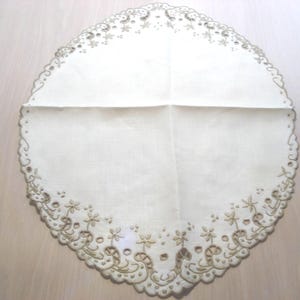 K&ouml;nnte beinhalten: Ovale, cremefarbene Tischdecke mit gewelltem Rand und gesticktem Blumendesign. Die Tischdecke hat einen detaillierten Rand mit ausgeschnittenen Mustern und kleinen Blumenmotiven, die dem Textil eine zarte Note verleihen.
