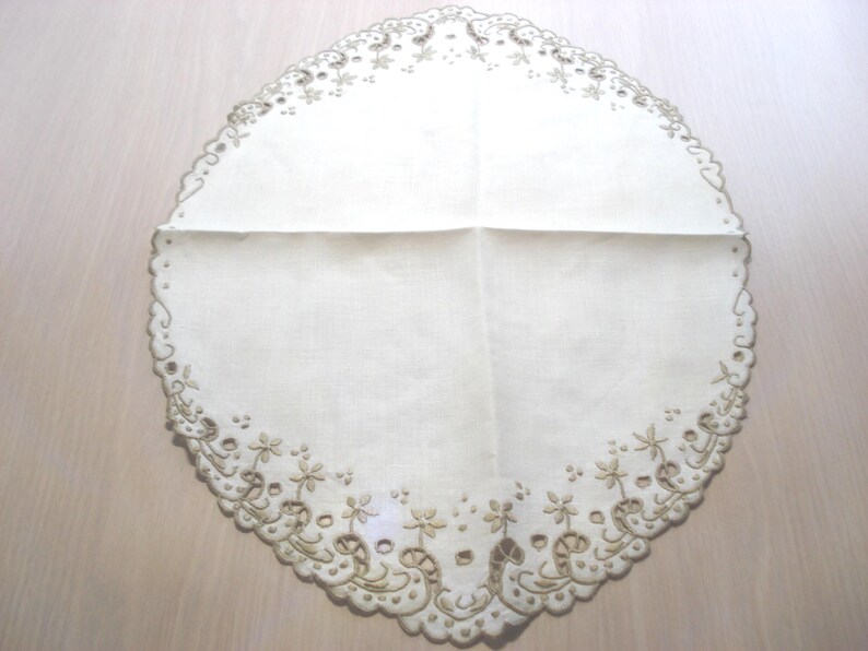 K&ouml;nnte beinhalten: Ovale, cremefarbene Tischdecke mit gewelltem Rand und gesticktem Blumendesign. Die Tischdecke hat einen detaillierten Rand mit ausgeschnittenen Mustern und kleinen Blumenmotiven, die dem Textil eine zarte Note verleihen.