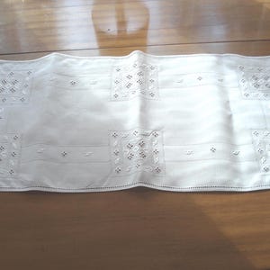 May include: White rectangular table runner with embroidered square and rectangular patterns. The runner is made of a soft, possibly linen, fabric. The embroidery features small, detailed designs, adding a touch of elegance to the piece. The runner is placed on a wooden table.