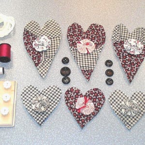 May include: A collection of sewing and craft supplies, including heart-shaped fabric ornaments, spools of thread, buttons, and embroidery floss. The hearts feature floral and gingham patterns in red, black, and white. Various sizes of buttons and spools are also visible.