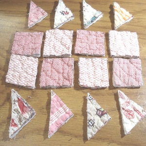 May include: An assortment of small, quilted fabric pieces in various shapes and patterns. The collection includes squares and triangles, featuring floral and geometric designs in shades of red, pink, and white. The pieces are arranged on a wooden surface.