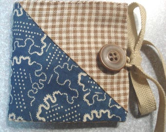 May include: A small, square fabric item with a brown and white checkered pattern and a blue patterned corner. It features a large, round brown button and a tan ribbon closure. The item appears to be handmade and has a homespun aesthetic.