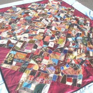 May include: A colorful, handmade quilt with a patchwork design. The quilt features a variety of fabric pieces in different colors and patterns, including floral, geometric, and abstract designs. The quilt is bordered with a deep red fabric and is laid out on a light blue sheet.