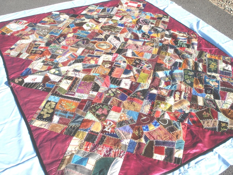 May include: A colorful, handmade quilt with a patchwork design. The quilt features a variety of fabric pieces in different colors and patterns, including floral, geometric, and abstract designs. The quilt is bordered with a deep red fabric and is laid out on a light blue sheet.
