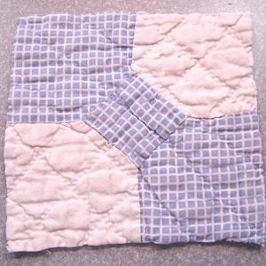 May include: Square quilted fabric piece with a patchwork design. The piece is divided into four sections, with two sections featuring a blue and white grid pattern and the other two sections featuring a white quilted pattern. The piece is likely a part of a larger quilt or decorative item.