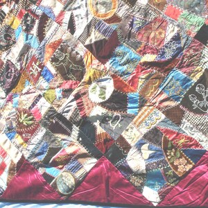 May include: A colorful patchwork quilt with a deep red border. The quilt features a variety of fabrics in shades of blue, red, brown, and green, with embroidered details and geometric patterns. The quilt's dimensions are not visible.