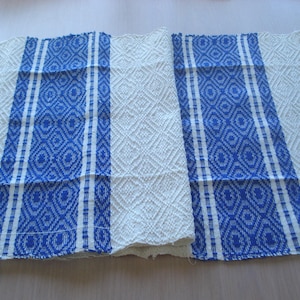 Op de afbeelding: Een wit en blauw geweven textiel met een ruitpatroon en twee verticale blauwe strepen. De strepen hebben een geometrisch ontwerp. Het textiel is waarschijnlijk een tafelloper of decoratieve doek.