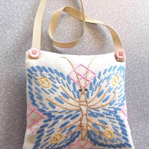 May include: A small, square, off-white fabric bag with a stitched butterfly design in blue, pink, and gold thread. The bag has a beige strap and is secured with two pink buttons. The butterfly design is the focal point.