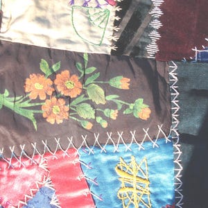 May include: Close-up of a patchwork textile with various fabric squares. The central square features a floral design with orange flowers and green leaves. The fabrics are in shades of pink, blue, brown, and black, stitched together with white thread.