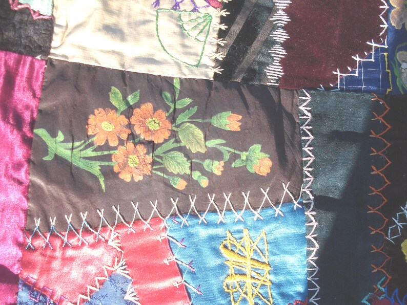 May include: Close-up of a patchwork textile with various fabric squares. The central square features a floral design with orange flowers and green leaves. The fabrics are in shades of pink, blue, brown, and black, stitched together with white thread.