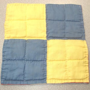 May include: A square, quilted fabric item with a patchwork design. The design features four equal squares, alternating between yellow and blue. The stitching is visible, and the edges are finished with a red border.