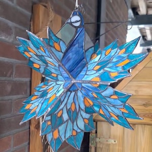 Könnte beinhalten: Ein Buntglas-Sonnenfänger in Form eines Pfaus mit blauen, türkisfarbenen und orangefarbenen Glasscheiben. Der Pfau hängt an einer Kette.