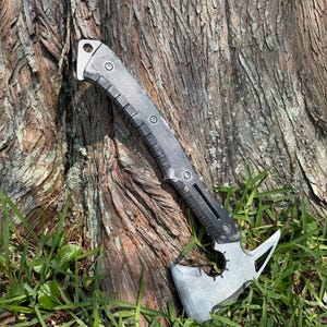 Pode incluir: Um machado tático tomahawk prateado e preto com um cabo texturizado e uma ponta no lado oposto da lâmina. O machado está apoiado na grama verde ao lado de um tronco de árvore.