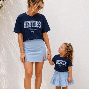 May include: A woman and a young girl wearing matching navy blue t-shirts with the word "BESTIES" printed on them. The woman wears a light blue skirt and white sneakers, while the girl wears a light blue tiered skirt and white sneakers. The text "Emma" and "Sophia" are printed on the shirts.