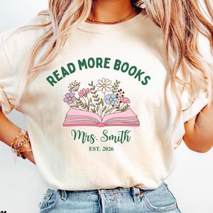 May include: An ivory t-shirt featuring a graphic of an open pink book with colorful flowers emerging from the pages. Above the book, the text reads "READ MORE BOOKS" in green. Below the book, the text reads "Mrs. Smith" and "EST. 2026".