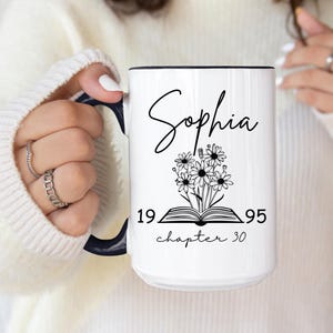 May include: White ceramic mug with a black design. The design features a book with flowers on top, the text "Sophia", and the numbers "19 95" with the text "chapter 30" underneath.