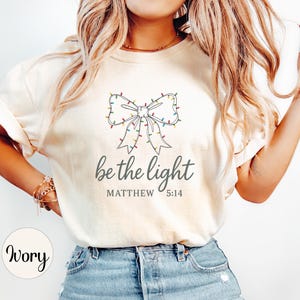 May include: Ivory-colored t-shirt featuring a bow design adorned with colorful Christmas lights. The text "be the light" is printed below the bow, with "MATTHEW 5:14" underneath. The shirt is made of a soft material.