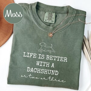 May include: A moss green t-shirt with the slogan "LIFE IS BETTER WITH A DACHSHUND or two or three" in white. The shirt features a dachshund graphic. A silver necklace with a round pendant rests on the shirt.
