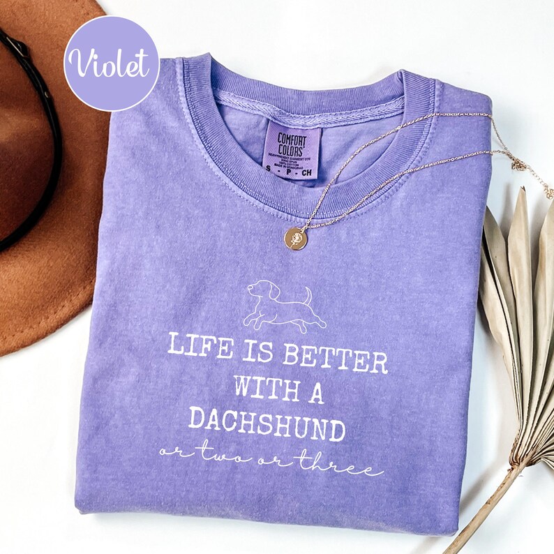 May include: A lilac Comfort Colors t-shirt with the text "LIFE IS BETTER WITH A DACHSHUND or two or three" in white. A gold necklace with a pendant is draped across the shirt. A brown hat and dried plant are in the background.