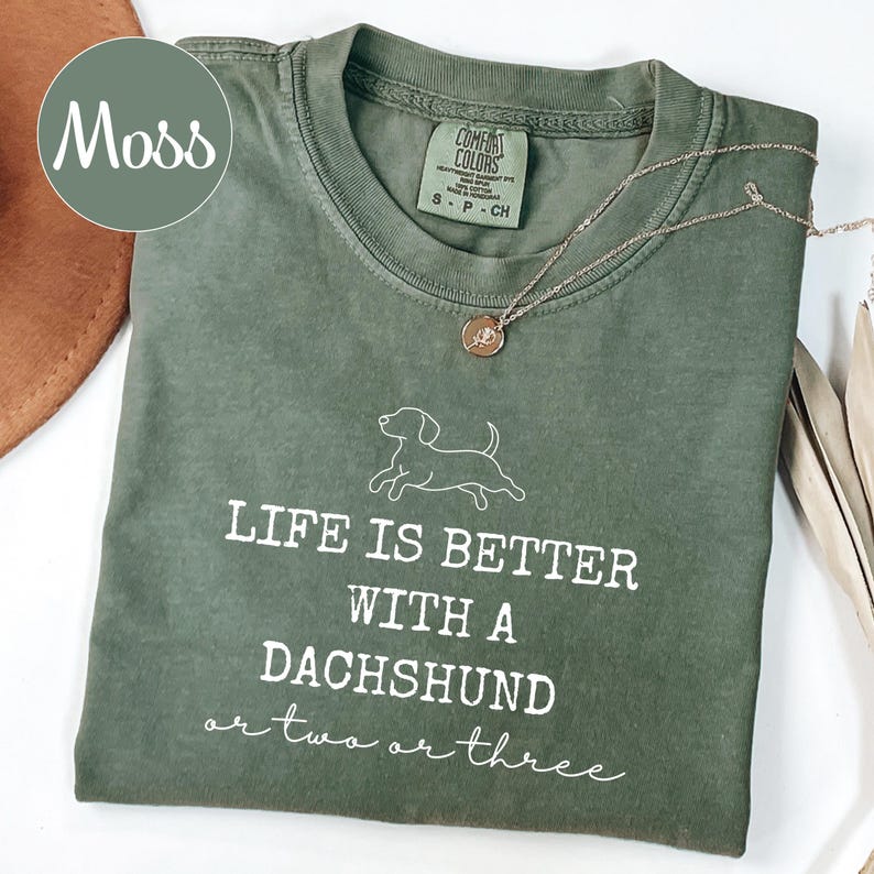 May include: A moss green t-shirt with the slogan "LIFE IS BETTER WITH A DACHSHUND or two or three" in white. The shirt features a dachshund graphic. A silver necklace with a round pendant rests on the shirt.