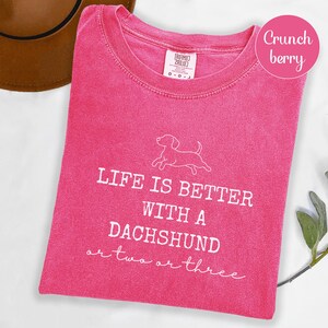 May include: A vibrant pink t-shirt featuring the white text "LIFE IS BETTER WITH A DACHSHUND or two or three" and a dachshund outline. The shirt has a crew neck and short sleeves. The colour is described as "Crunch berry".
