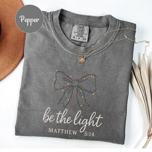 May include: A gray Comfort Colors t-shirt with a bow design made of Christmas lights. The text "be the light" and "Matthew 5:14" are printed below the bow. A gold necklace is draped across the shirt.