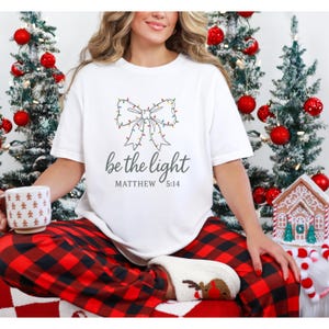 May include: White t-shirt with a bow design made of Christmas lights and the text "be the light" with "Matthew 5:14" underneath. The person is wearing red and black plaid pajama pants and reindeer-themed socks. Christmas trees and decorations are in the background.