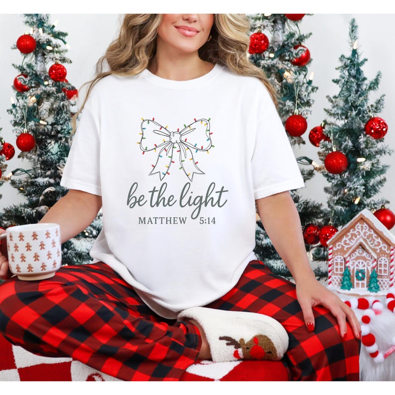 May include: White t-shirt with a bow design made of Christmas lights and the text "be the light" with "Matthew 5:14" underneath. The person is wearing red and black plaid pajama pants and reindeer-themed socks. Christmas trees and decorations are in the background.