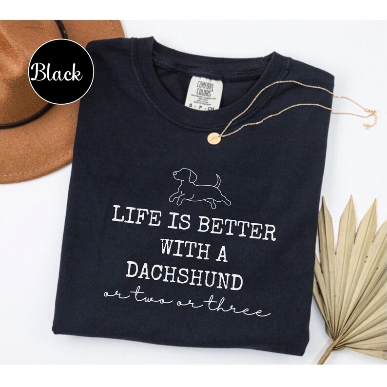 May include: Black crew neck t-shirt with the text "LIFE IS BETTER WITH A DACHSHUND or two or three" and a dachshund illustration. A gold necklace and a brown hat are also visible.