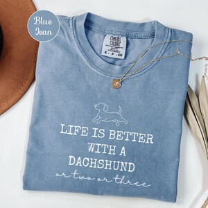 May include: A blue jean Comfort Colors t-shirt with the text "LIFE IS BETTER WITH A DACHSHUND or two or three" in white. A gold necklace with a pendant is on the shirt. The shirt is folded and placed on a white surface.