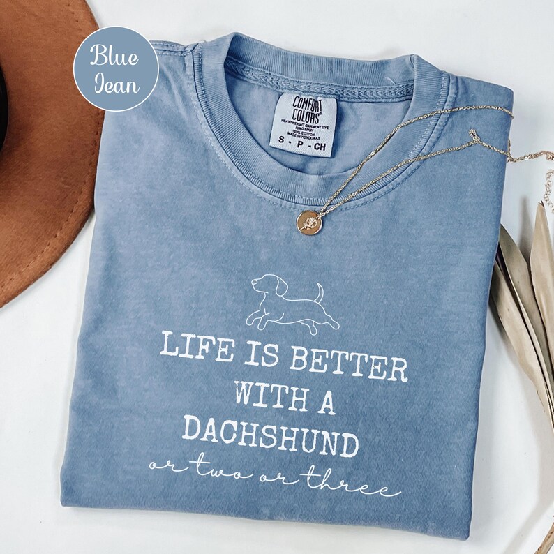 May include: A blue jean Comfort Colors t-shirt with the text "LIFE IS BETTER WITH A DACHSHUND or two or three" in white. A gold necklace with a pendant is on the shirt. The shirt is folded and placed on a white surface.