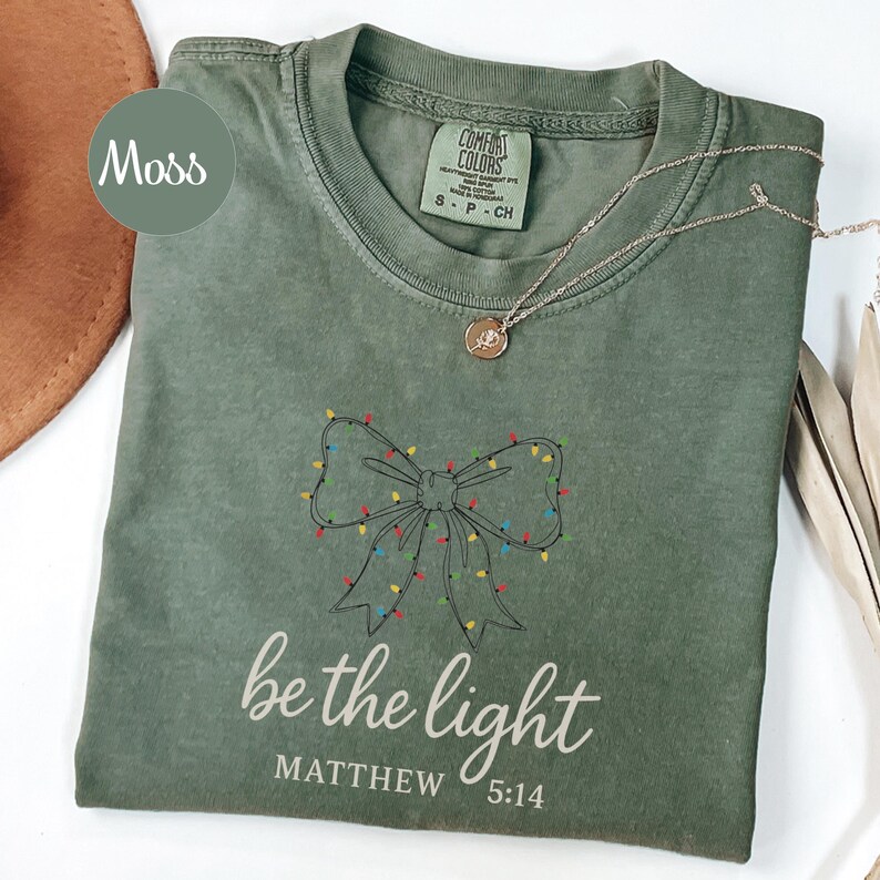 May include: A moss green t-shirt with a bow design made of Christmas lights. The text "be the light" and "Matthew 5:14" are printed below the bow. A silver necklace with a pendant is on the shirt.