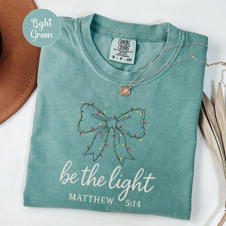 May include: Light green t-shirt with a bow design made of Christmas lights. The text "be the light" and "Matthew 5:14" are printed in white. A silver necklace with a pendant is draped on the shirt.