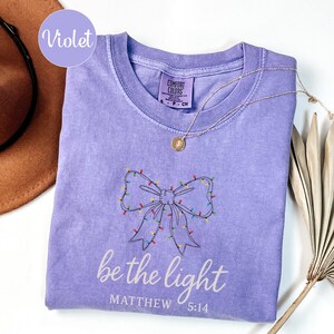 May include: Violet-colored Comfort Colors t-shirt with a bow design made of Christmas lights. The text "be the light" and Matthew 5:14 are printed below the bow. A gold necklace with a pendant is on the shirt.