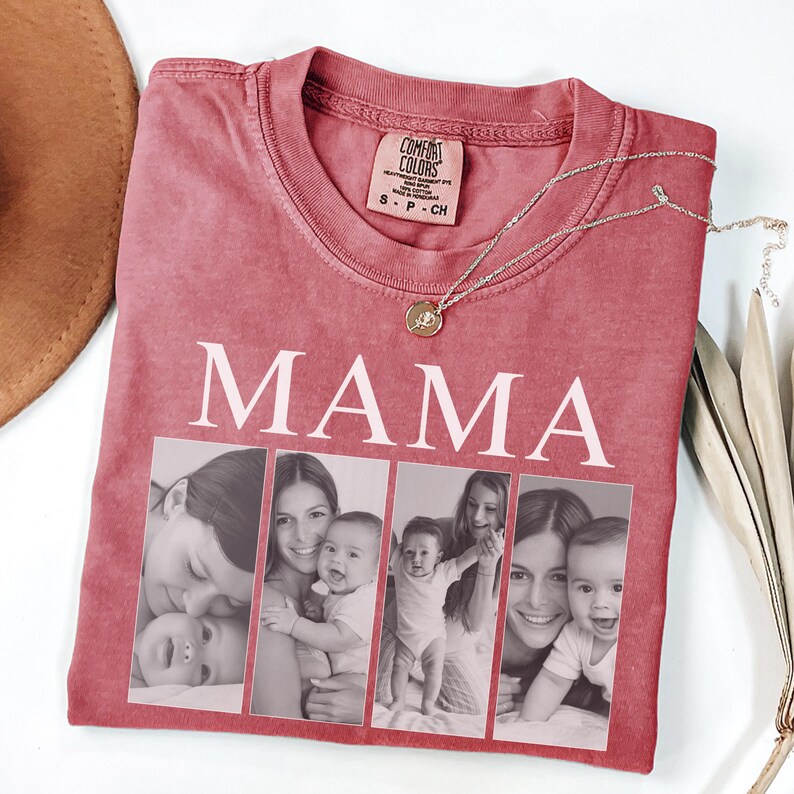 May include: A dusty rose t-shirt featuring the word MAMA in white capital letters above four vertical rectangular photo frames. Each frame contains a black and white photograph of a person holding a baby. A gold chain necklace rests on the shirt.