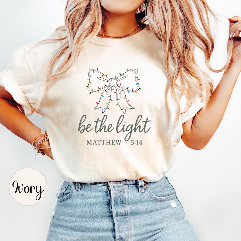 May include: Ivory-colored t-shirt featuring a bow design adorned with colorful Christmas lights. The text "be the light" is printed below the bow, with "MATTHEW 5:14" underneath. The shirt is made of a soft material.