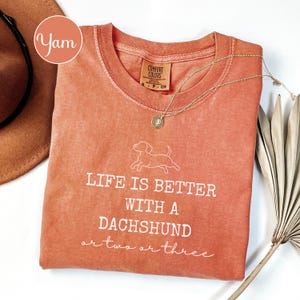 May include: A coral-coloured t-shirt with the phrase "LIFE IS BETTER WITH A DACHSHUND or two or three" in white script. A gold necklace with a pendant and a dachshund graphic are also visible.
